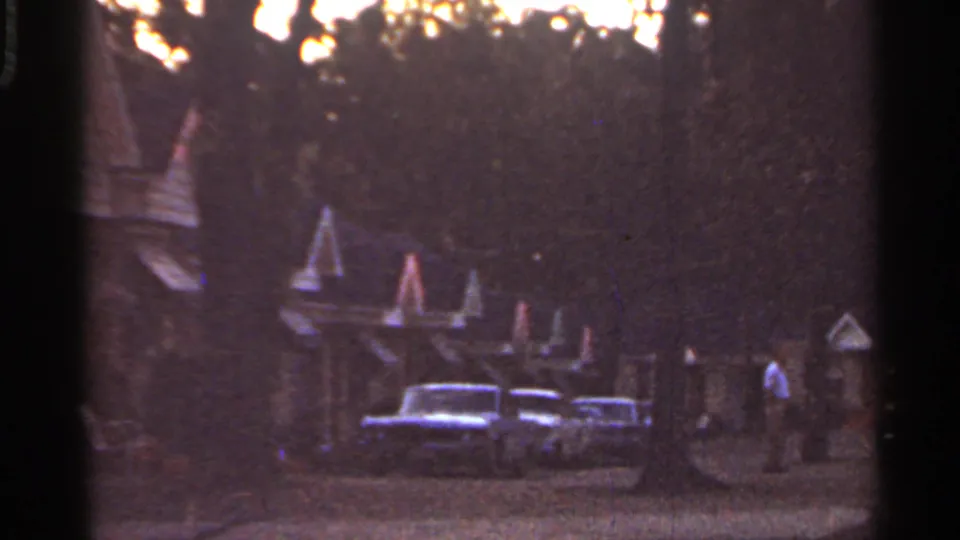 1965 Arkansas 8mm frame showing a darker motel frontage with vintage cars, peaked roof forms, trees, and a person at right.