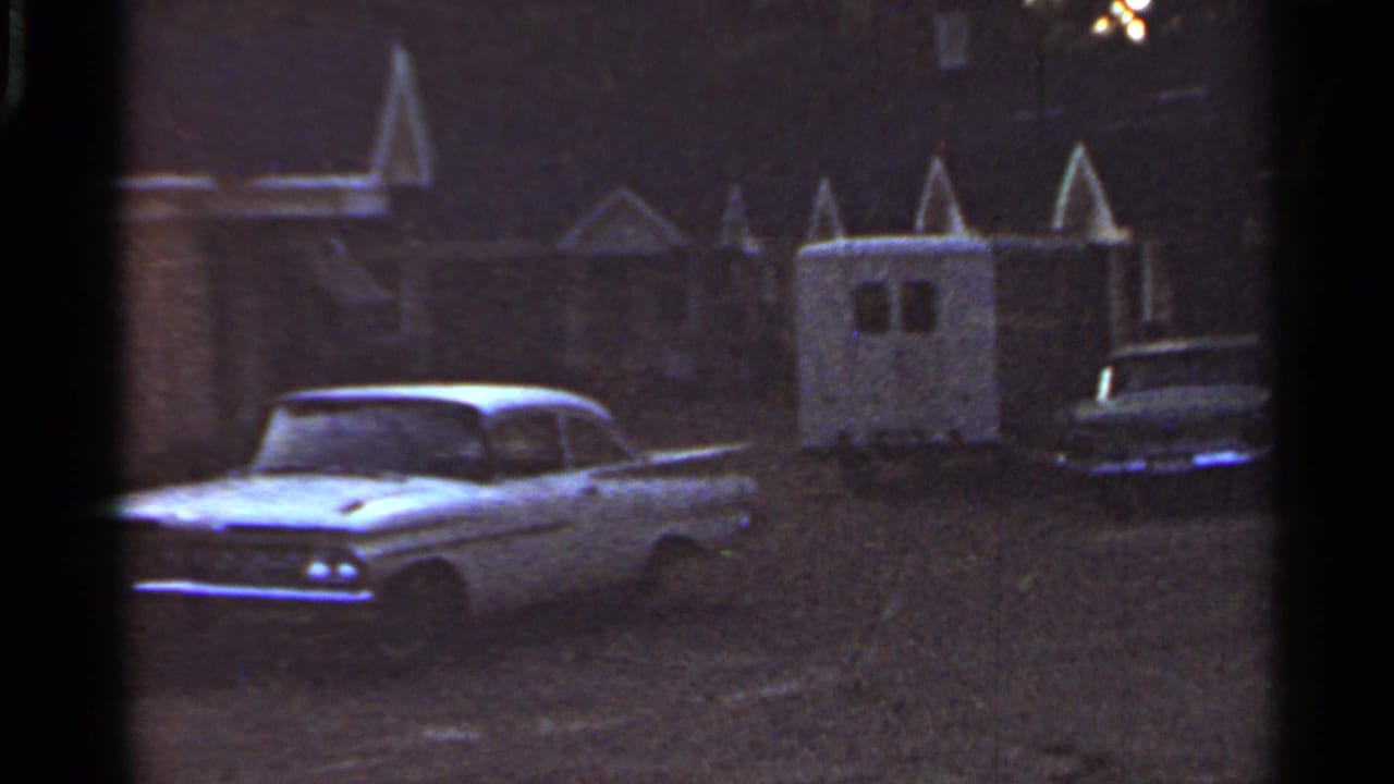 1965 Arkansas home-movie frame with a white car, small trailer, and quiet suburban exteriors in low light.