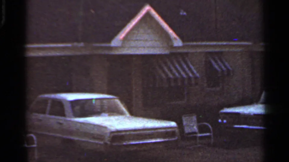 1965 Arkansas home-movie frame of a parked white car, house awnings, and a dark car in a quiet yard.