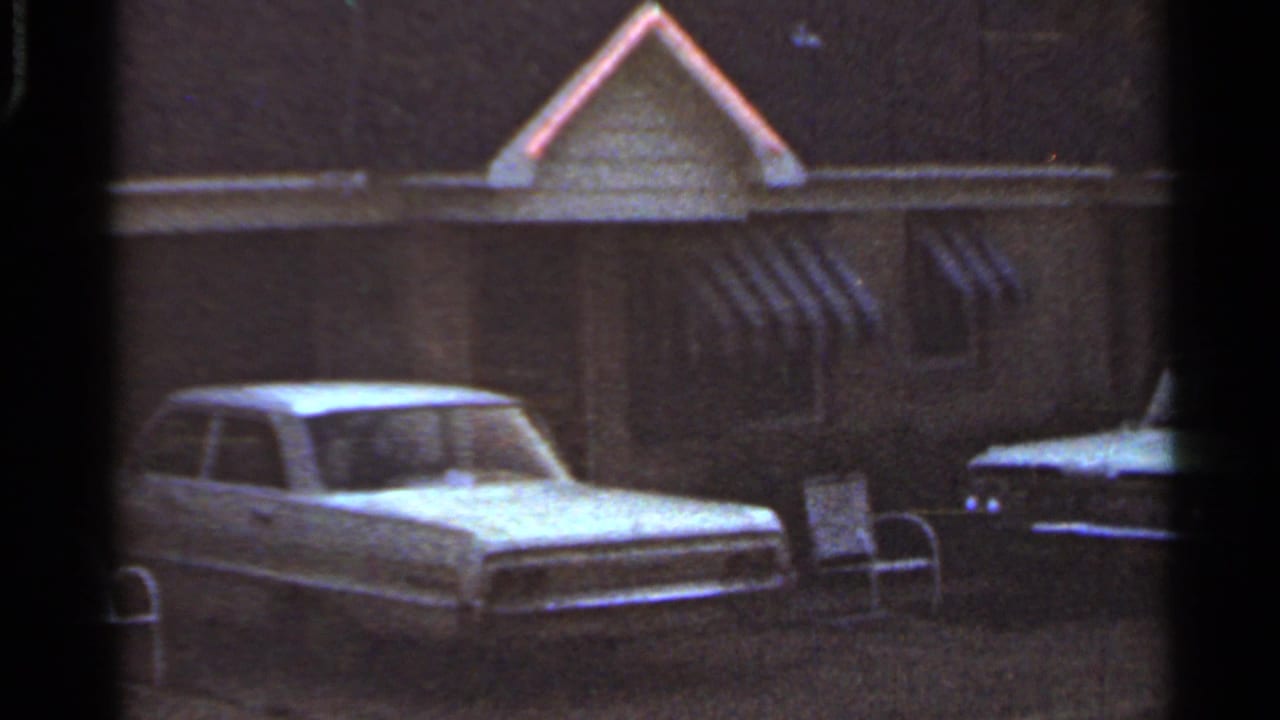 1965 Arkansas 8mm frame showing a parked car near a modest exterior with striped awnings and a dark vehicle at the edge of frame.