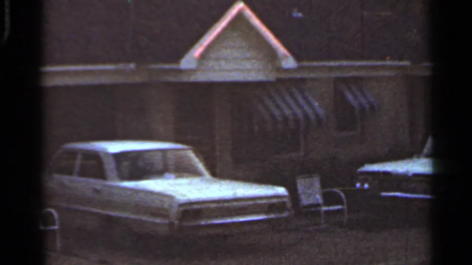 1965 Arkansas 8mm frame showing a parked car near a modest exterior with striped awnings and a dark vehicle at the edge of frame.