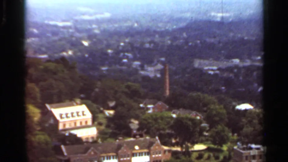 1965 Arkansas frame showing a hillside town scene with brick buildings, houses, trees, and a slender tower.