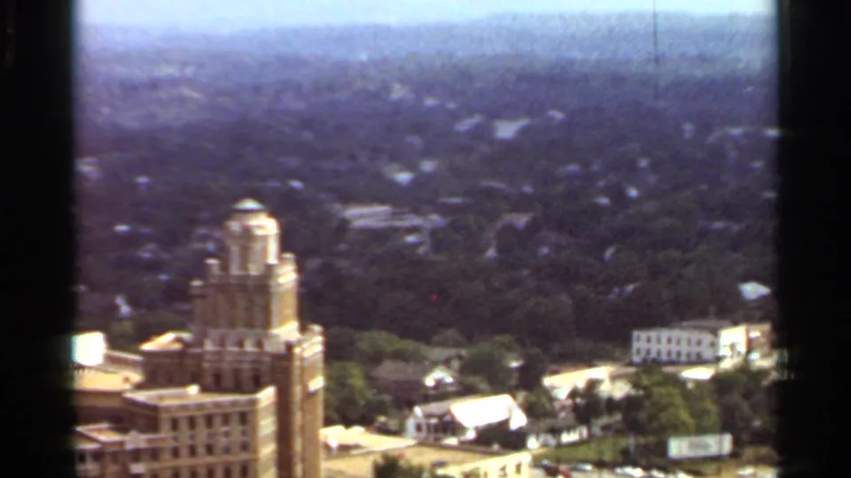 1965 Arkansas 8mm frame showing a city view with a distinctive tower, nearby buildings, trees, and hazy residential areas.