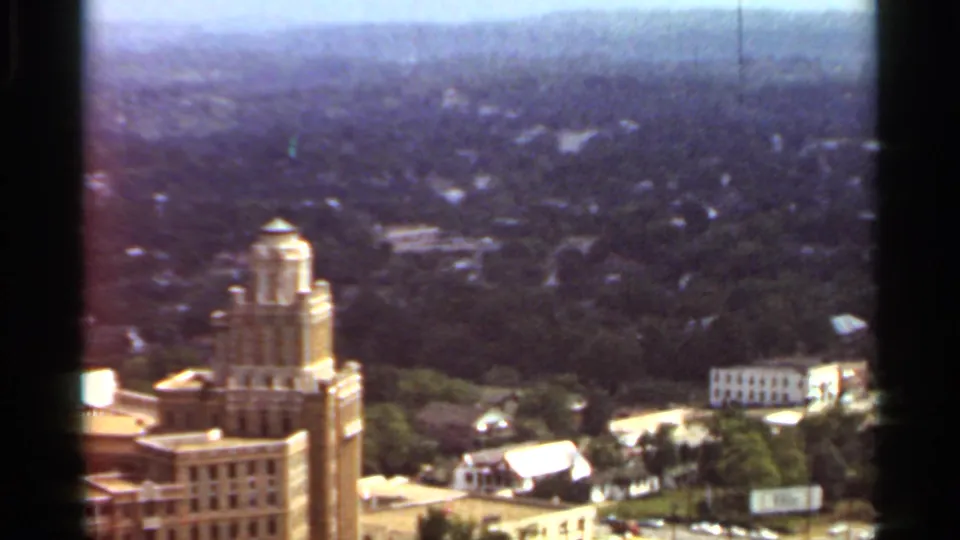 1965 Arkansas 8mm frame showing an elevated view over a city with a tall downtown building, tree canopy, roads, and low-rise structures.