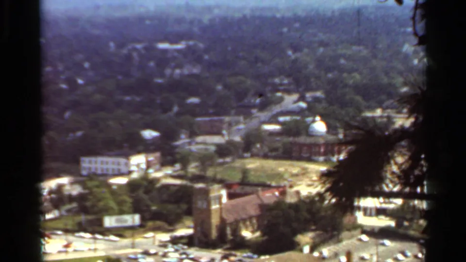 1965 Arkansas 8mm view over church towers, roadways, parked cars, scattered business buildings, and a leafy urban setting.