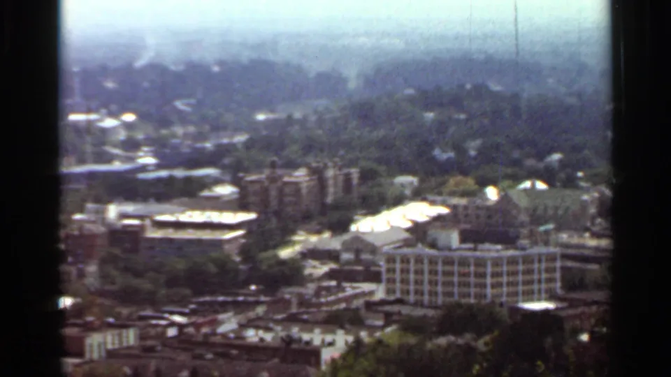 1965 Arkansas home-movie frame showing downtown rooftops, a taller building, roads, parking areas, and tree-covered neighborhoods from above.