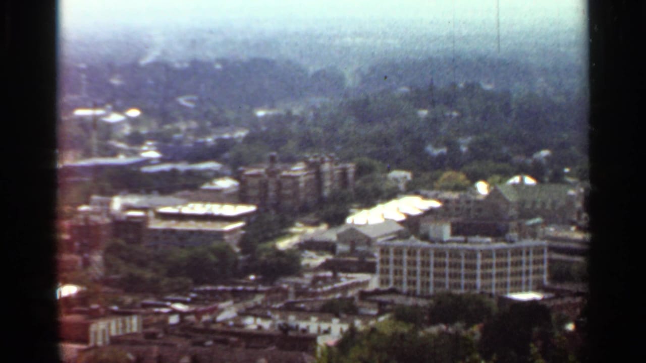 1965 Arkansas 8mm frame showing a hazy elevated view over mid-century buildings, rooftops, and tree cover.