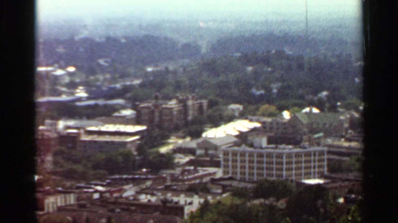 Similar elevated city view with clustered buildings, rooftops, trees, and pale sky.