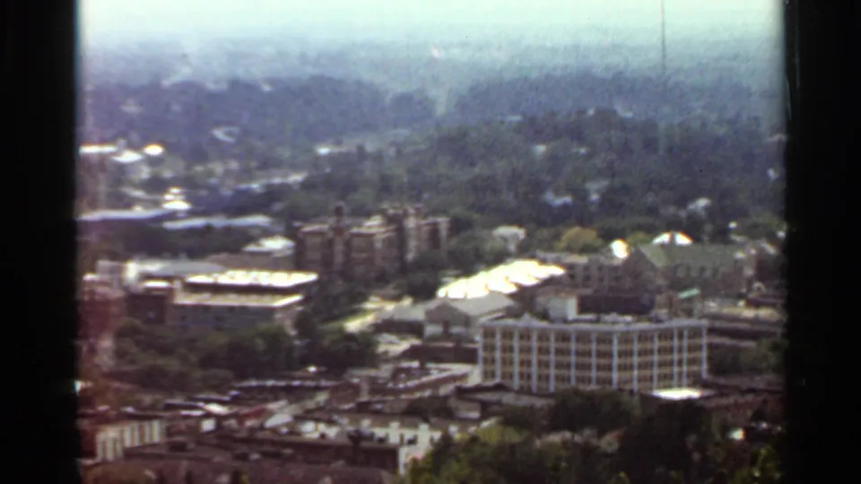1965 Arkansas home-movie frame with a high view of urban blocks, low rooftops, dense trees, and a muted sky.