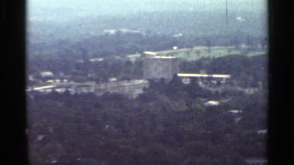 1965 Arkansas 8mm frame showing a distant rectangular building set among trees, roads, and hazy rooflines.