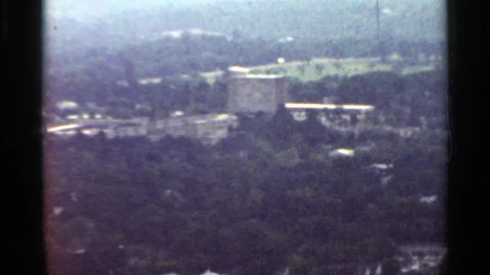 1965 Arkansas frame with a distant urban structure above dense trees, road traces, and faded 8mm color.