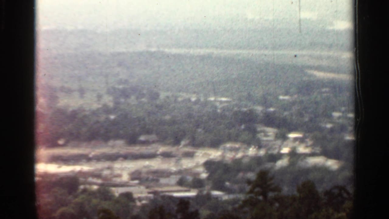 1965 Arkansas 8mm frame showing a hazy elevated view over trees, scattered buildings, pale roads, and distant low hills.