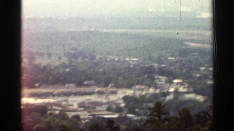 1965 Arkansas 8mm frame showing a hazy elevated view over trees, scattered buildings, pale roads, and distant low hills.