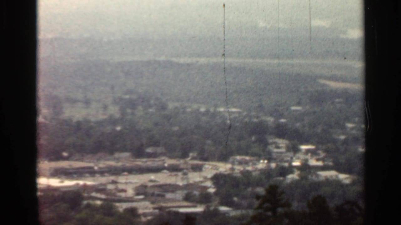 1965 Arkansas home-movie frame with a soft, elevated view across tree cover, town buildings, roads, and hazy hills.
