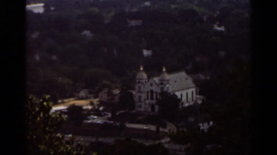 1965 Arkansas frame showing a white church with twin domes below a wooded hillside, seen through dark 8mm borders.