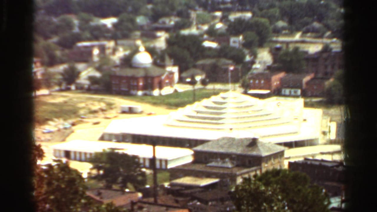 1965 Arkansas 8mm frame showing a tiered pyramid-like roof building, red brick domed building, roads, trees, and rooftops.