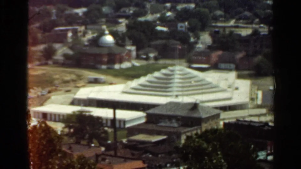 1965 Arkansas home-movie frame showing a stepped-roof modernist building near brick structures, trees, and a domed church.