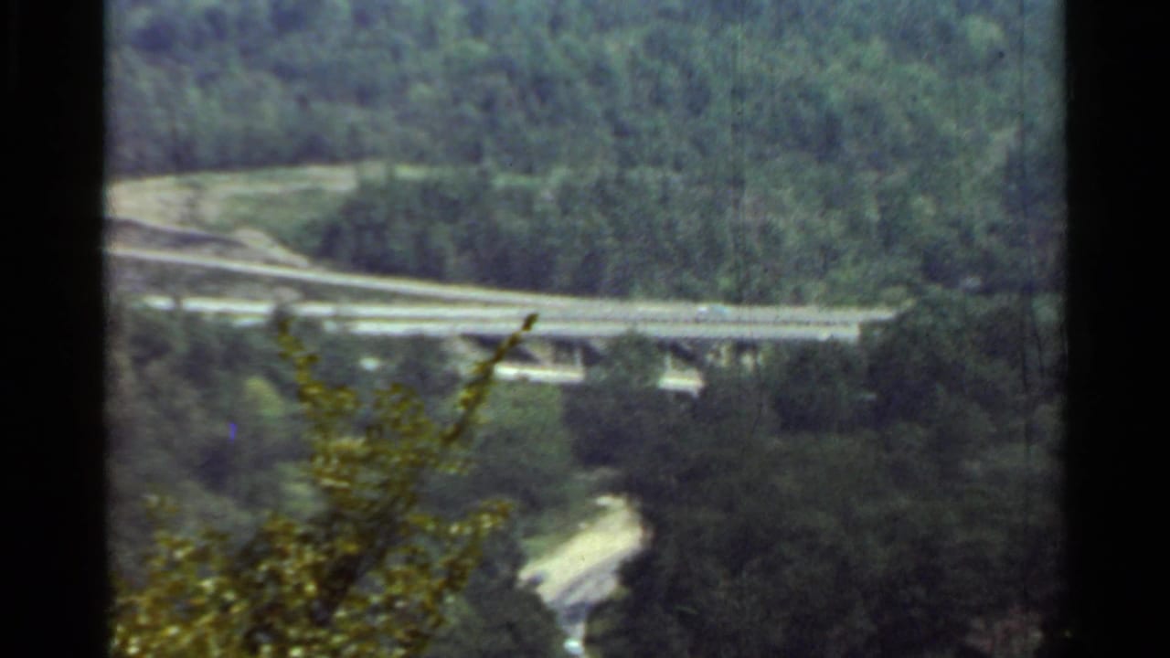 1965 Arkansas frame showing a highway overpass curving across a wooded valley with a lower winding road partly visible.
