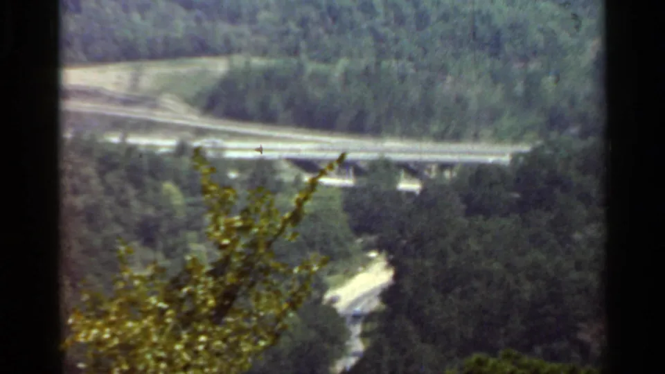1965 Arkansas frame showing a curving highway overpass, supporting piers, trees, and a pale road below in the valley.