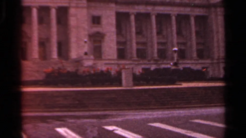 Arkansas 1965 home-movie frame showing a columned government-building facade across a road, with lawn and orange flowers.