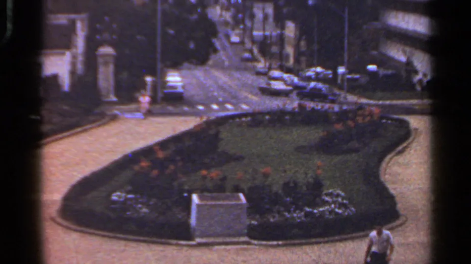 Wide 1965 Arkansas view of a shaped garden bed with flowers, hedges, a stone marker, parked cars, and buildings nearby.