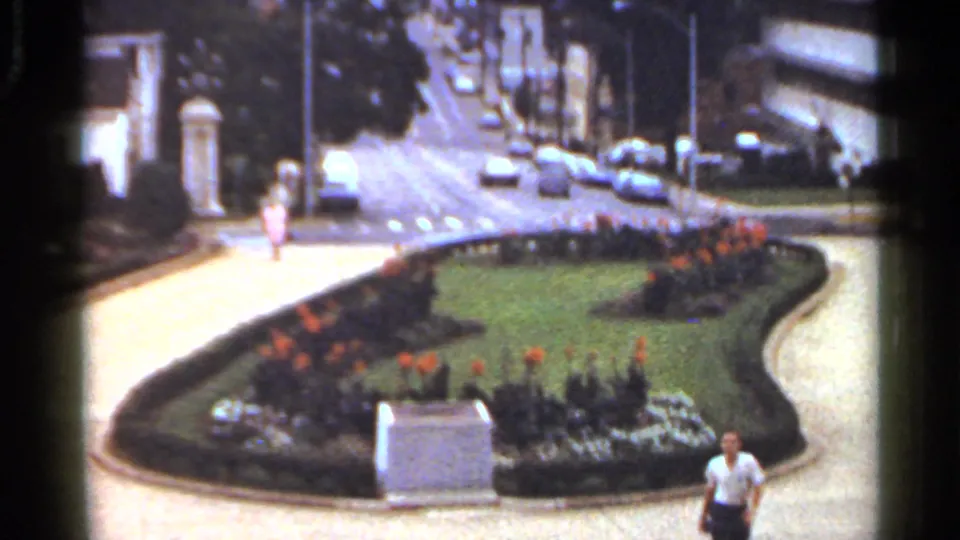 Wide shot of the Arkansas city garden with a shaped lawn, flower beds, stone marker, parked cars, and a man at the lower edge.