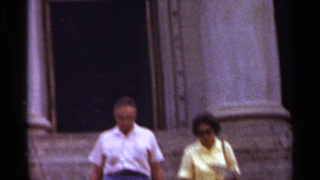Older couple, likely visitors, walking outside a monumental Arkansas building in 1965 home-movie footage.