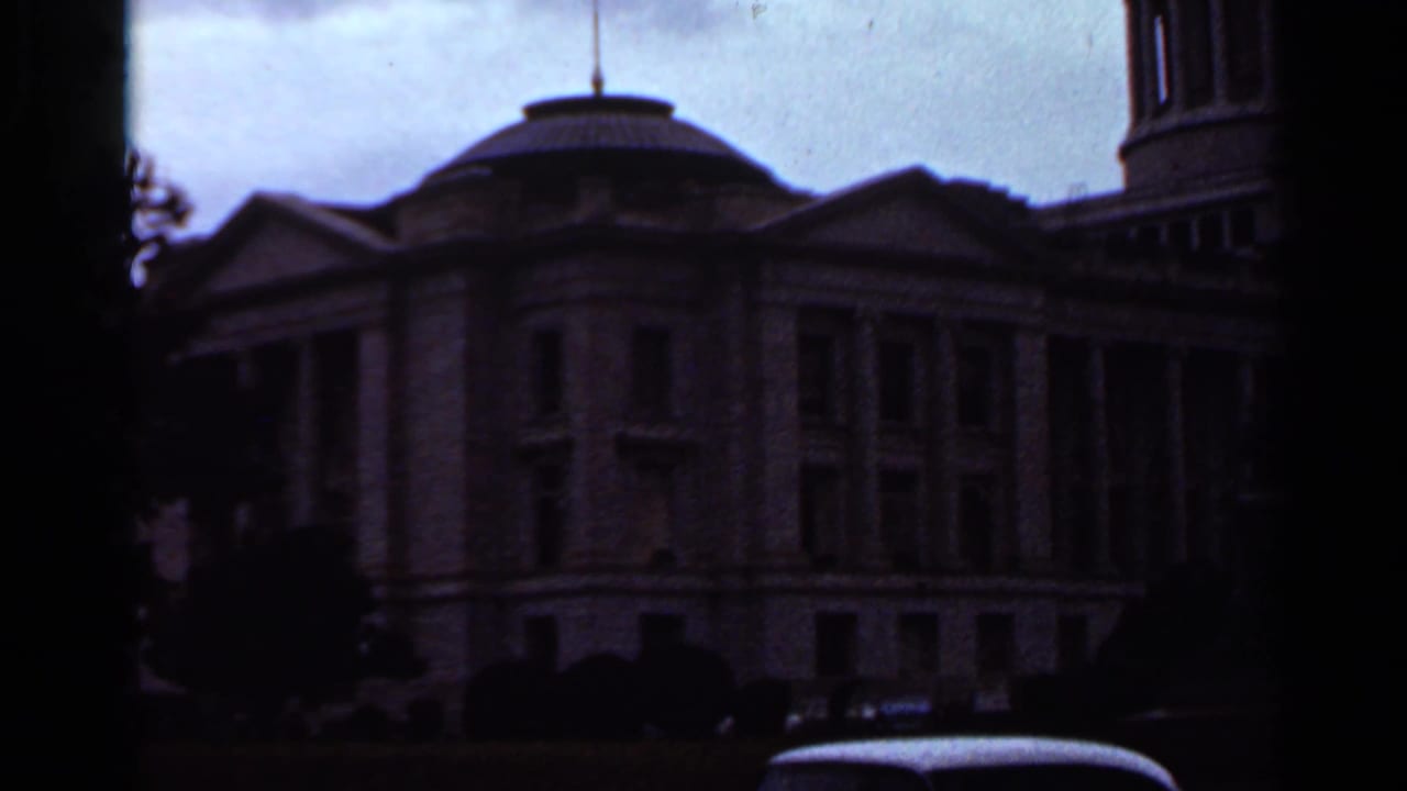 Underexposed 1965 8mm view of the Arkansas State Capitol in Little Rock, with the dome, columns, trees, and a car roof partly visible.