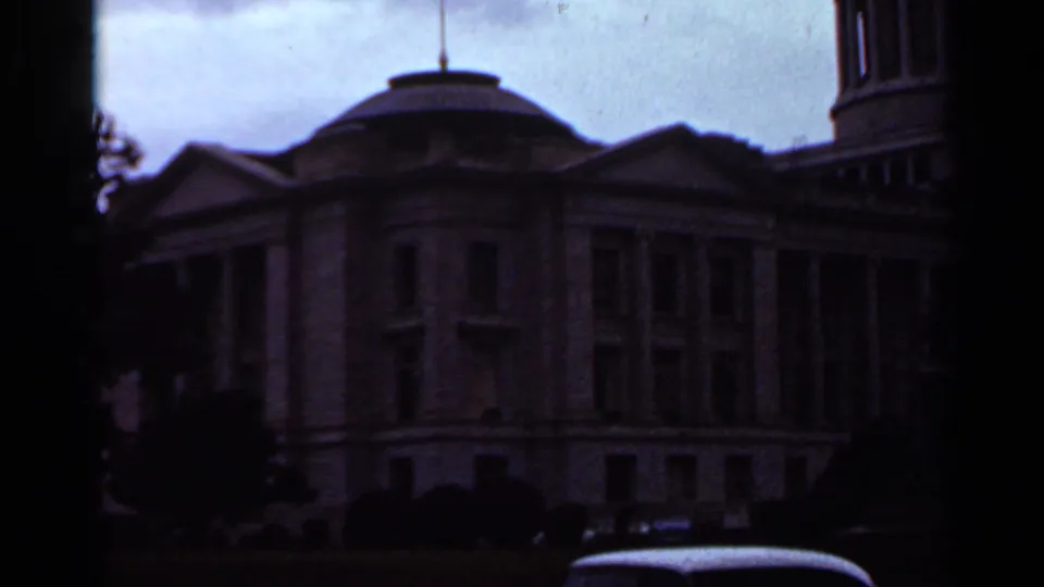 Underexposed 1965 8mm view of the Arkansas State Capitol in Little Rock, with the dome, columns, trees, and a car roof partly visible.