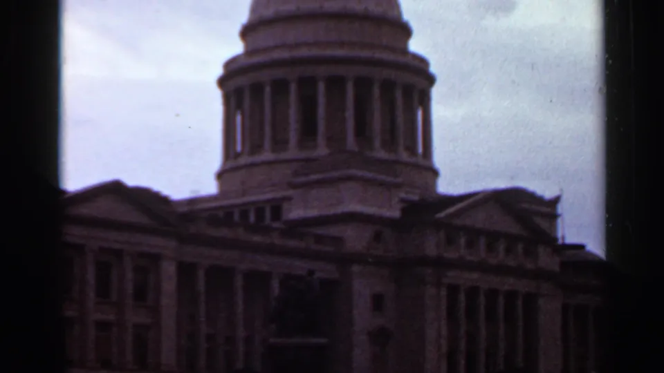 Visitor-shot 1965 8mm view of the Arkansas State Capitol dome, side wings, and columns, with grain and dark gate edges visible.