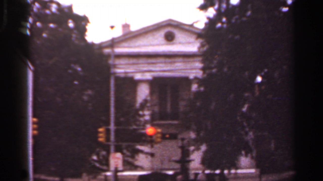 Columned building behind trees and a red traffic light, viewed from a moving vehicle.
