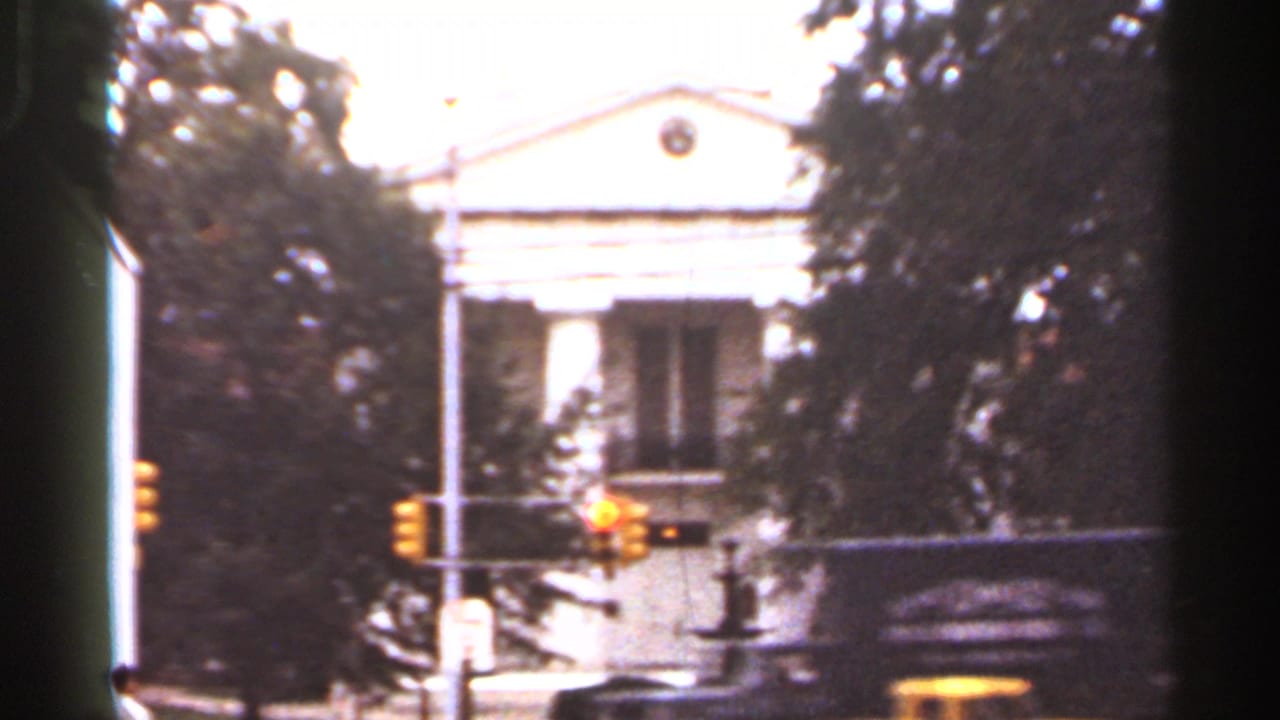 Bright view of a columned building, trees, traffic lights, and a bus or vehicle edge.