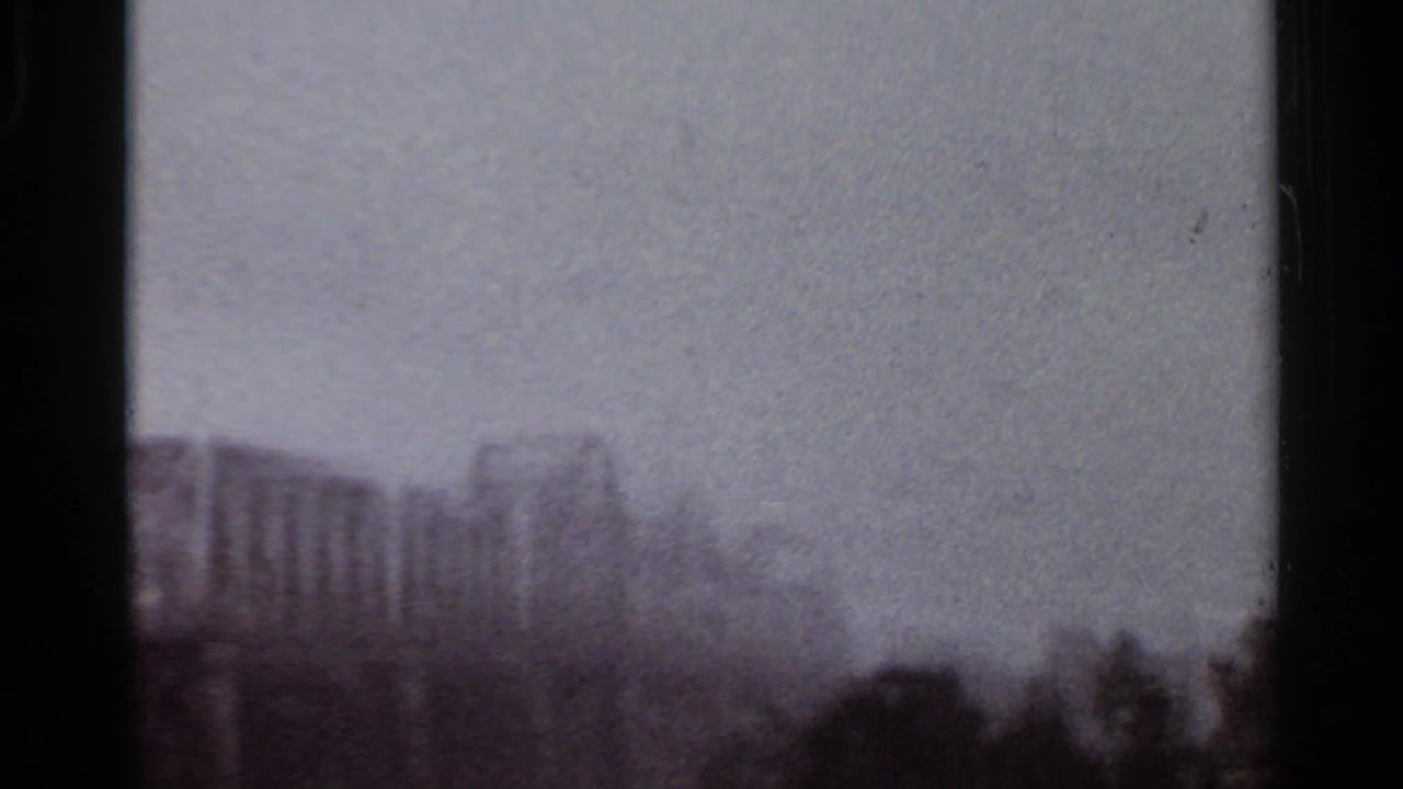 Rainy windshield view with hazy buildings and dark roadside shapes beyond the glass.