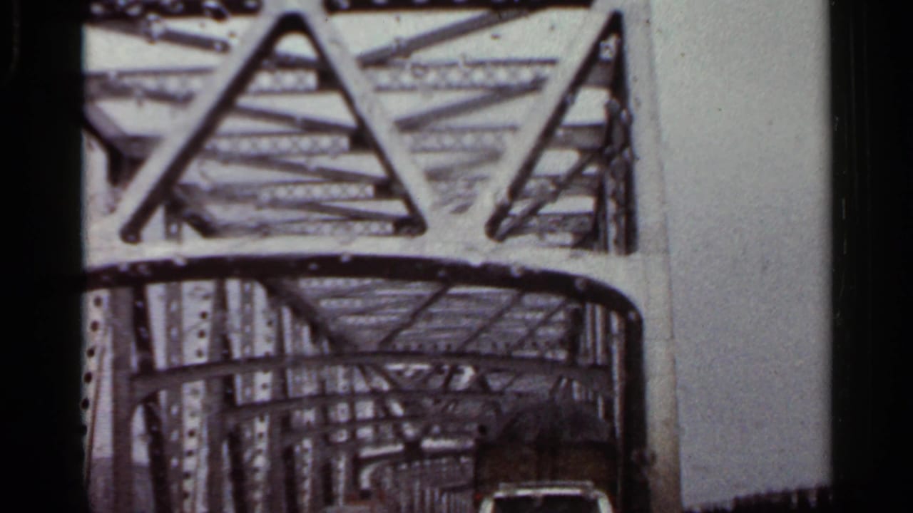Steel truss bridge seen from inside a moving vehicle, with overhead beams and a car ahead.