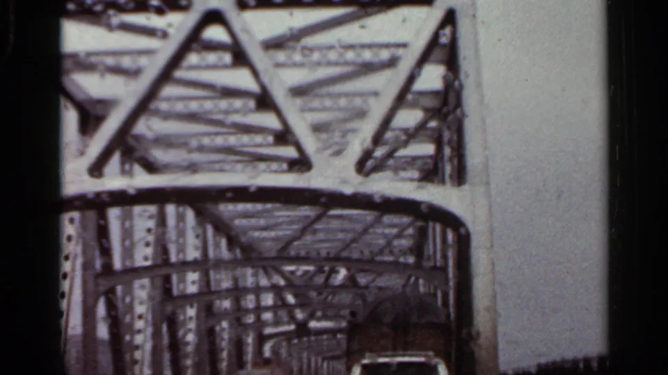 1965 Arkansas 8mm frame from inside a vehicle entering a steel truss bridge with overhead latticework and traffic ahead.
