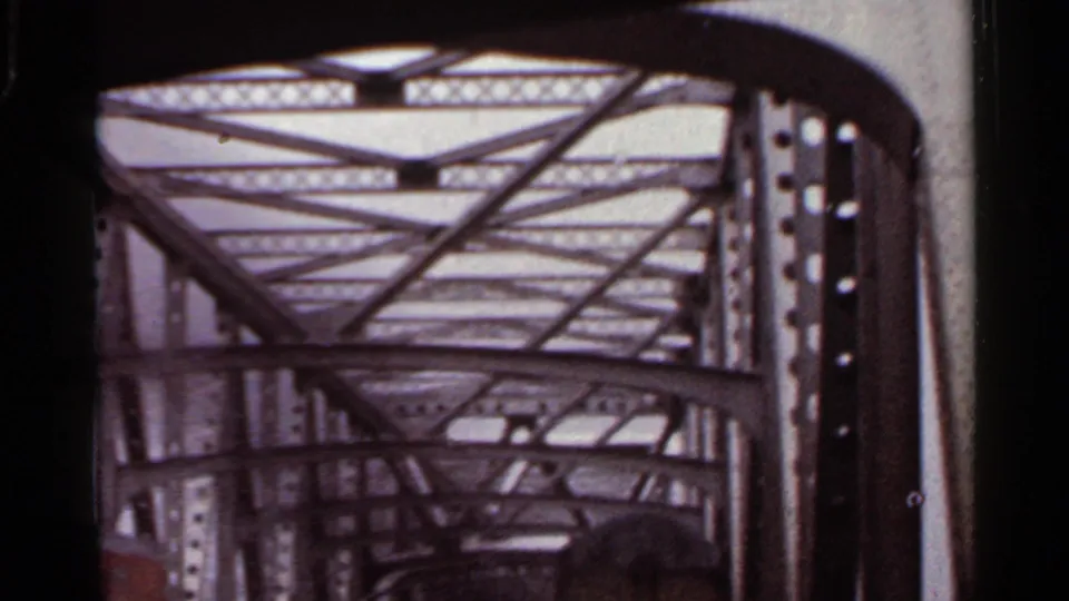 1965 Arkansas 8mm frame looking forward under a steel truss bridge, with repeating beams and a vehicle ahead in rainy light.