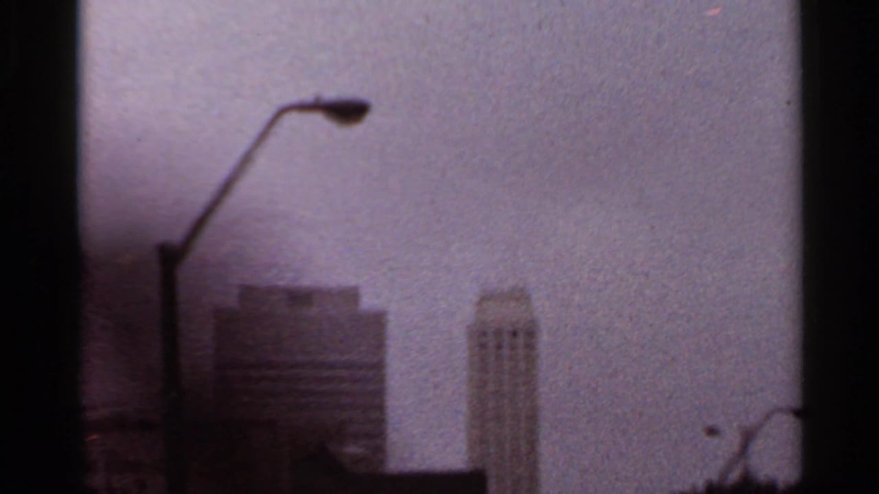 Rainy windshield view of a streetlamp and hazy tall buildings under an overcast sky.