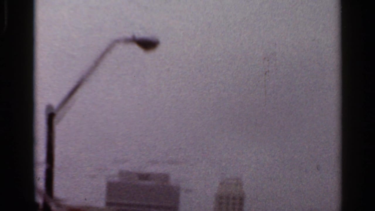Streetlamp in the foreground with blurred high-rise buildings beyond a wet windshield.
