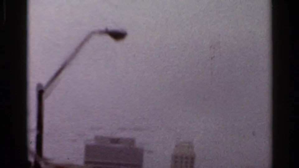 Rain-dimmed vehicle-window view of an Arkansas urban skyline, with a foreground streetlamp and soft 8mm grain.