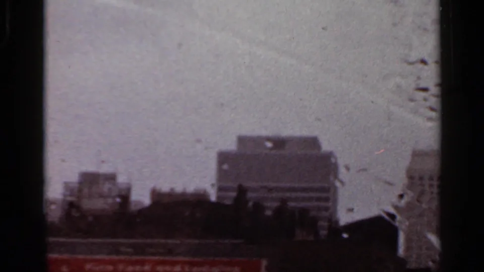 1965 Arkansas home-movie frame with low rooftops, larger downtown buildings, and water streaks on the vehicle glass.
