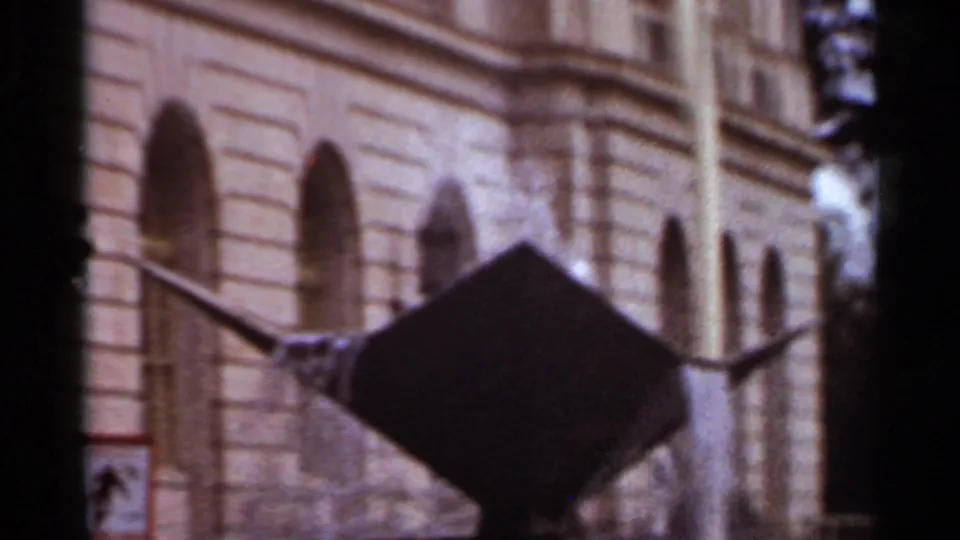 1965 Arkansas 8mm frame centers the dark metal fountain against a tan arched facade, with trees near the edge.