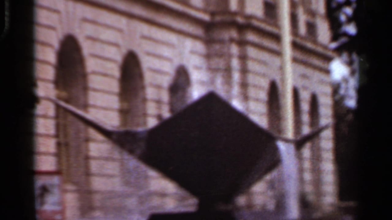 1965 Arkansas 8mm frame showing a stone building with arched windows and a dark angular sculpture or sign in front.