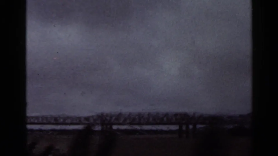 1965 Arkansas home-movie frame with a steel bridge, wide water, low shore, and tiny distant figures near the crossing.