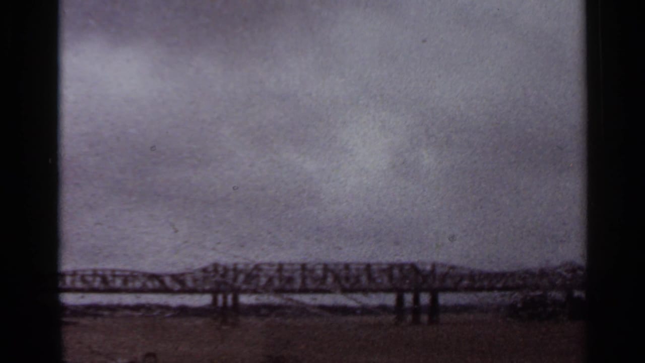 1965 Arkansas home-movie view of a distant steel truss bridge under heavy cloud cover, framed by dark film edges.