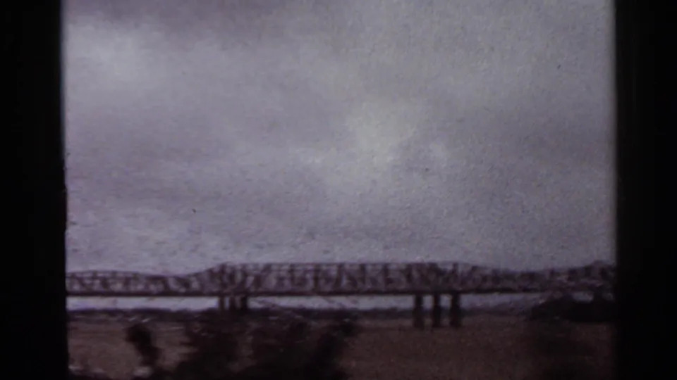 Distant Arkansas steel truss bridge in 1965 home-movie footage, with gray sky and low waterway visible below.