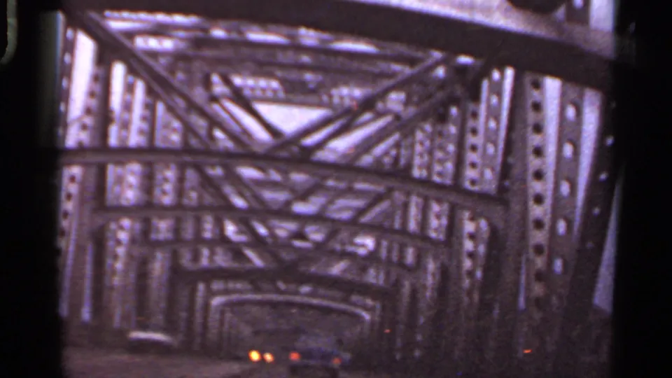1965 Arkansas car-window view inside a steel truss bridge, with repeating beams, riveted uprights, and dim lights ahead.