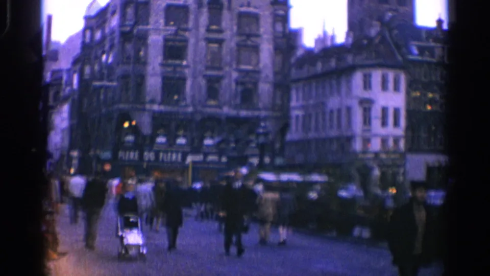 1965 Copenhagen street view with pedestrians crossing or walking near storefronts, parked cars, and a pram in soft 8mm color.
