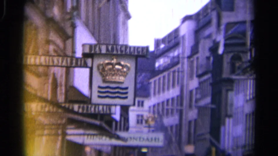 1965 Copenhagen view centered on the crowned Da Konge sign, with layered building facades, storefront awnings, and visible 8mm color shift.