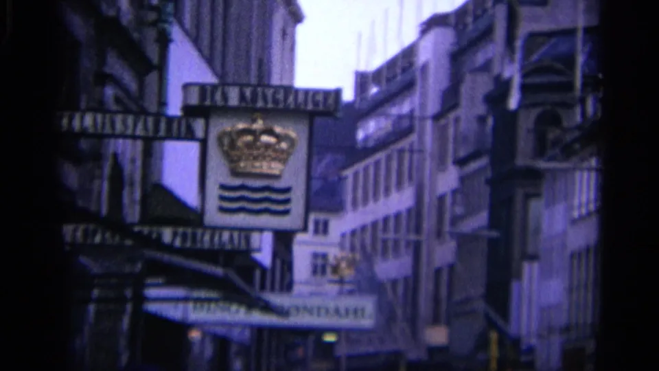 1965 Copenhagen street view with a crown-and-waves hanging sign, commercial facades, awnings, and pedestrians below.