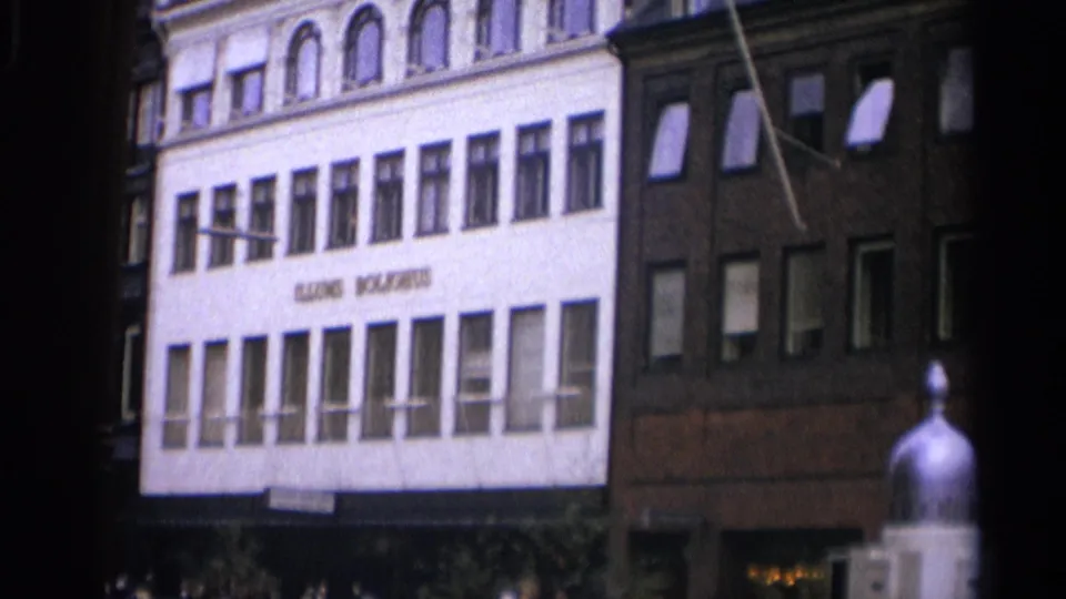1965 Copenhagen frame showing a white commercial facade with visible lettering, a neighboring brick building, windows, awnings, and pedestrians below.
